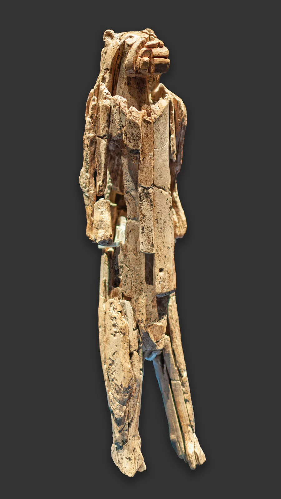 A pale ivory figure standing upright against a dark grey background. It has a lion's head and a long, mostly human body with arms at its sides. The surface is cracked and patched where many fragments have been fitted together.