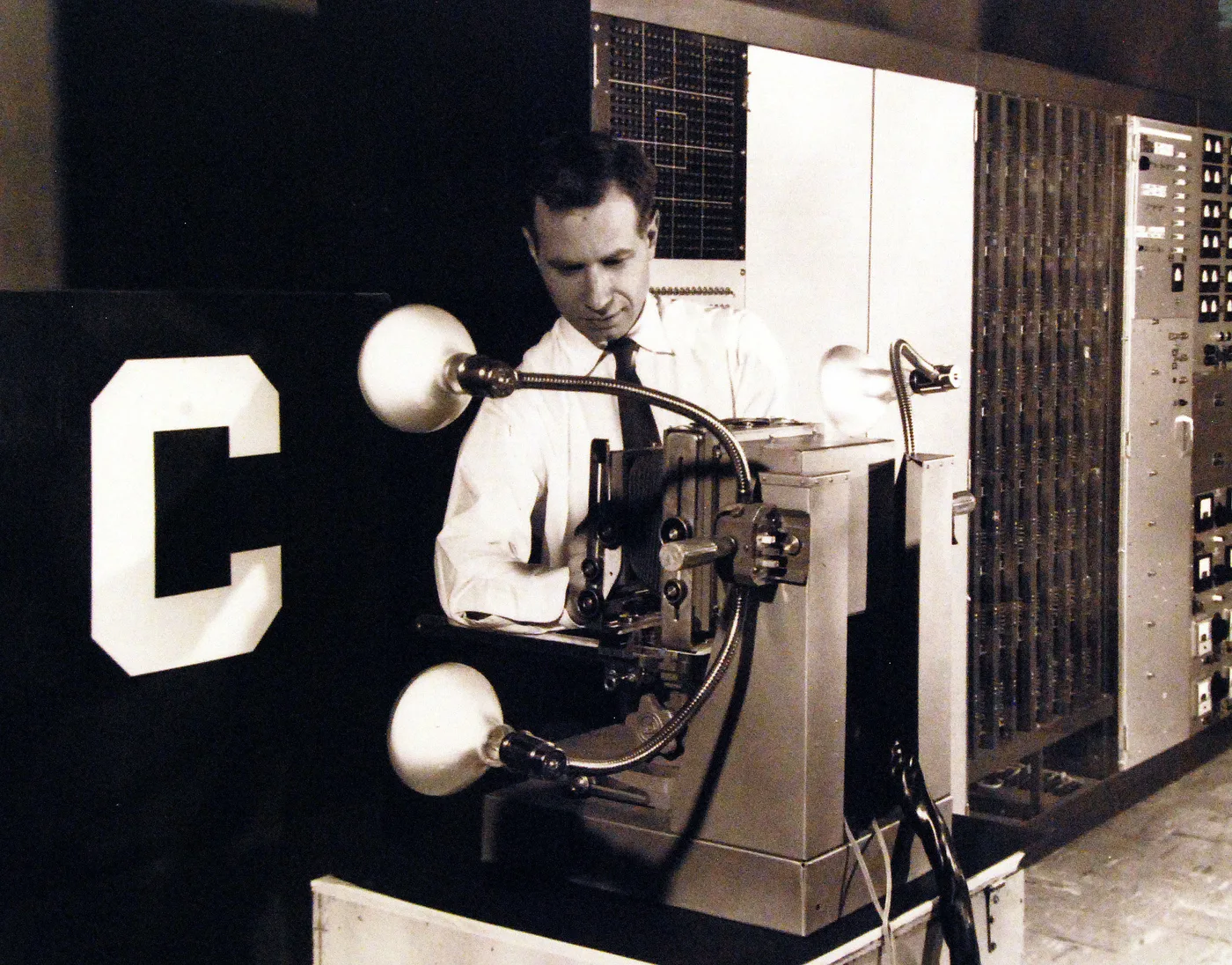 A black-and-white photograph. A man in a white shirt and tie works at a camera-like unit on a stand, lit by three lamps on flexible arms. In front of it, a large white letter C on a black card faces the lens. Racks of wiring and a control panel fill the wall behind.